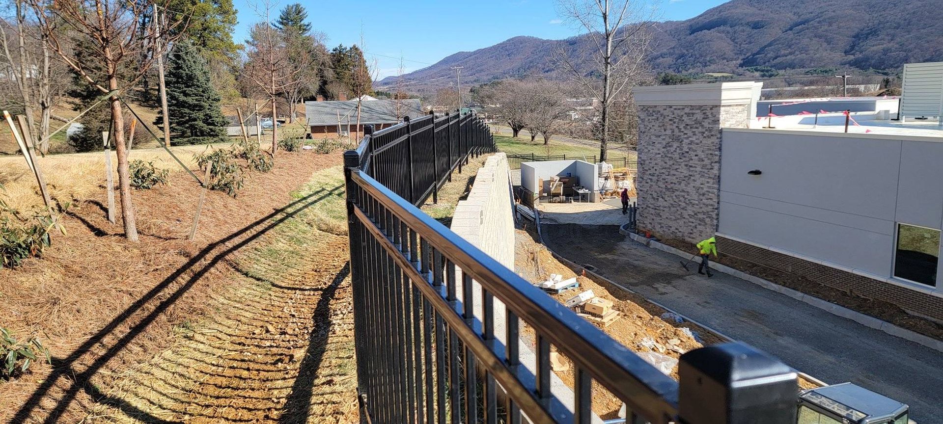 A black metal fence curves along a brown landscape, with a building on the right and mountains in the background.
