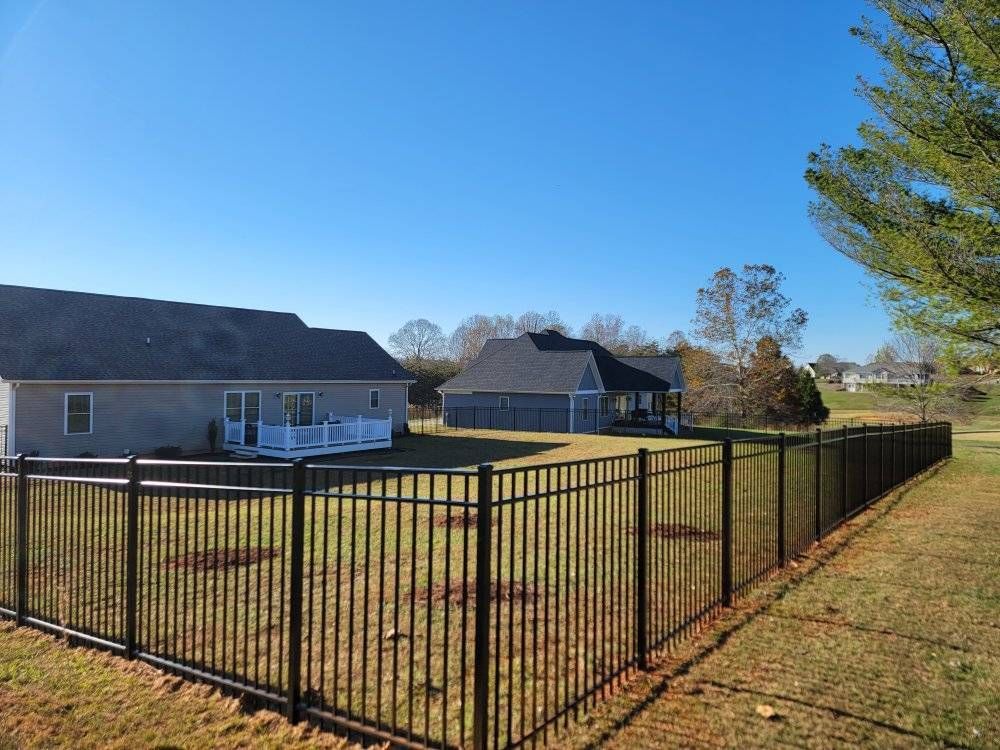 Black metal fence surrounds grassy yard with two houses under a bright blue sky.