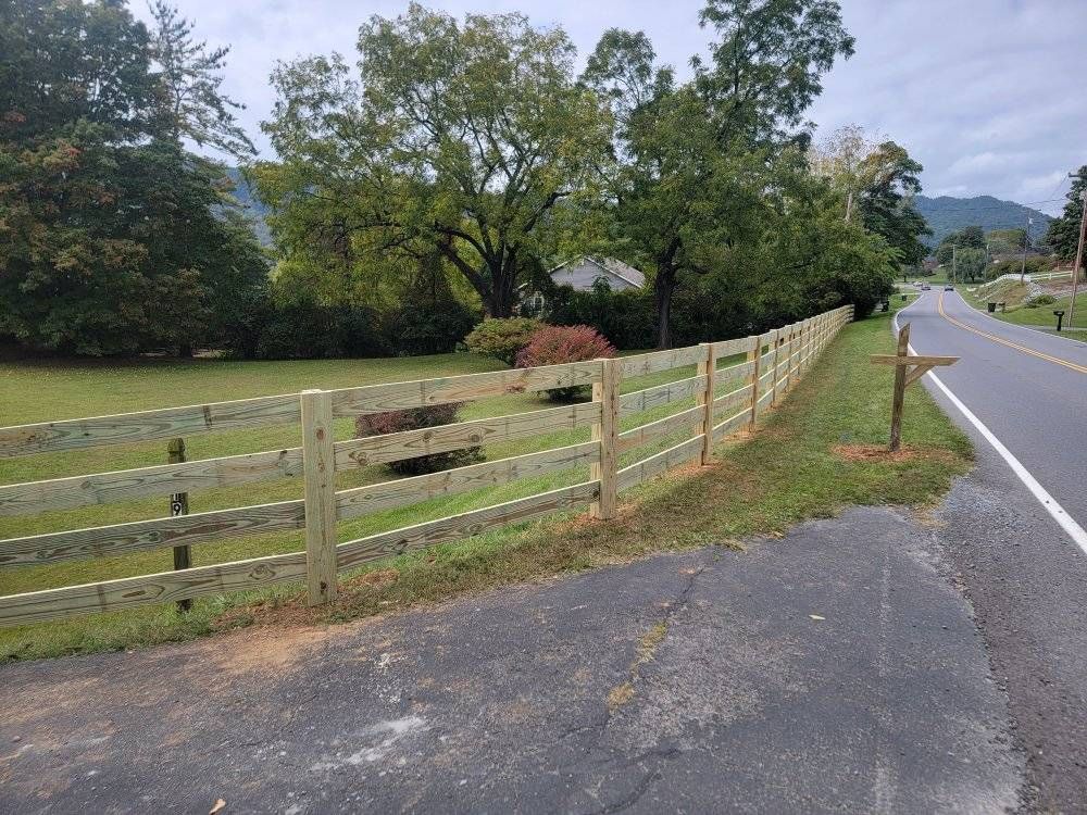 Wooden split-rail fence alongside a road, separating a grassy area from the pavement. Trees in background.