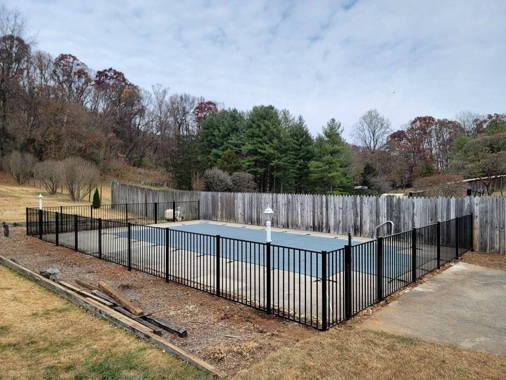 A rectangular outdoor swimming pool surrounded by a black fence and wooden wall, set in a yard with fall foliage and a cloudy sky.