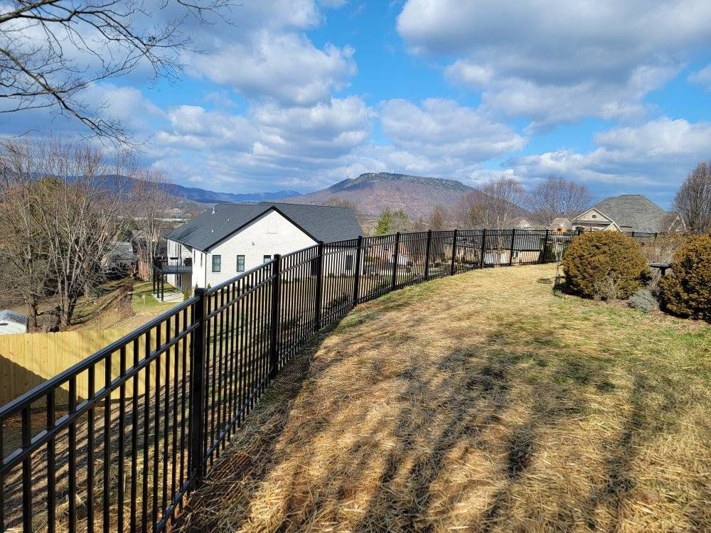 Black metal fence on a hillside, overlooking mountains and houses under a partly cloudy sky.
