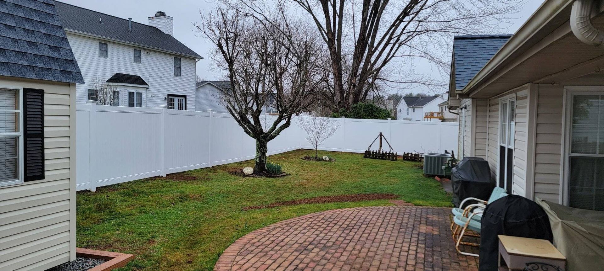 A backyard with a brick patio, green grass, a bare tree, a white fence, and houses on a cloudy day.