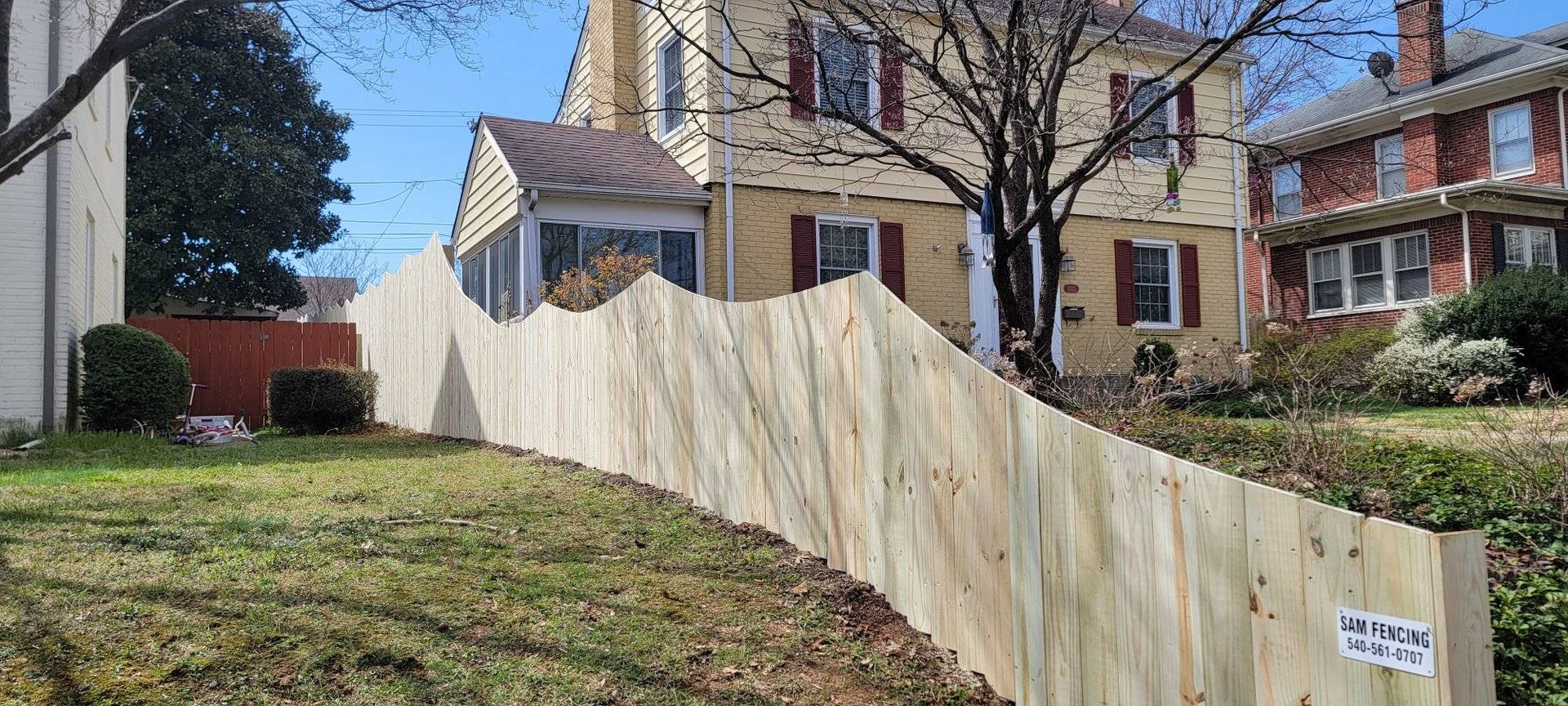 A light-colored wooden fence curves along a grassy yard, in front of a yellow house with red shutters.