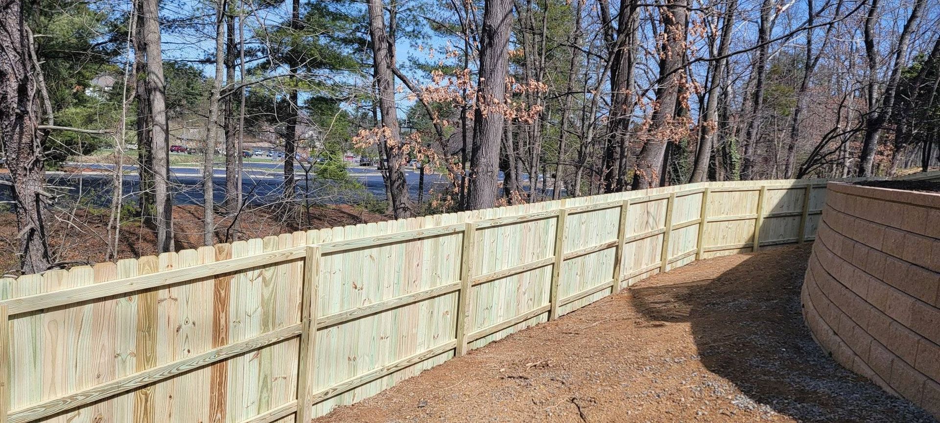 Wooden fence curves along a brown, graveled yard, with trees and a road visible in the background.