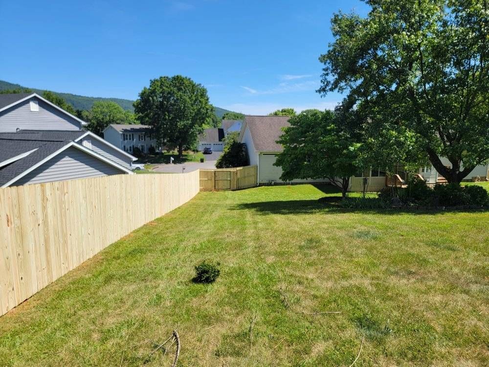 Wooden fence encloses a green lawn, with houses and a tree in the background under a blue sky.