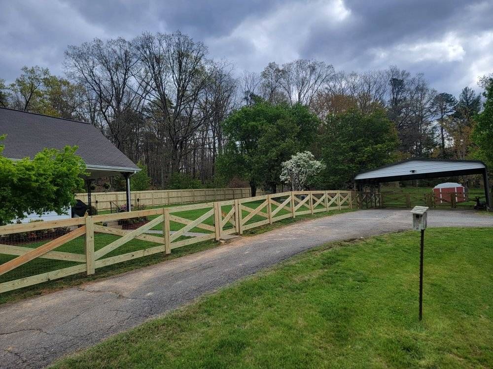 Wooden fence along a driveway with trees in the background under a cloudy sky.
