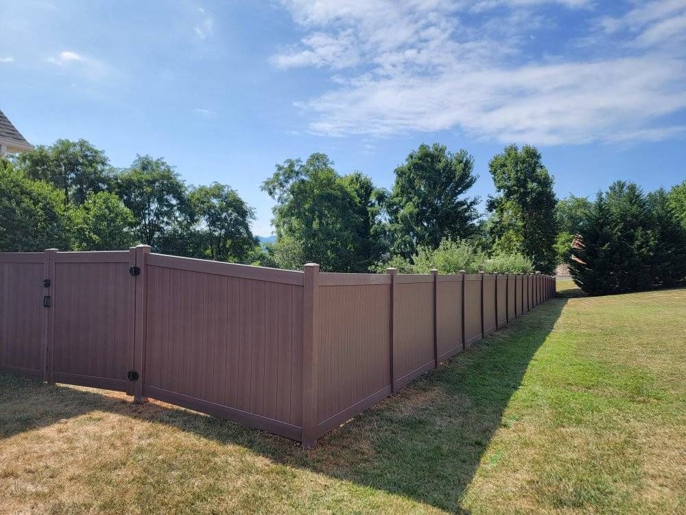 Brown vinyl fence in a grassy yard, with trees and a blue sky in the background.
