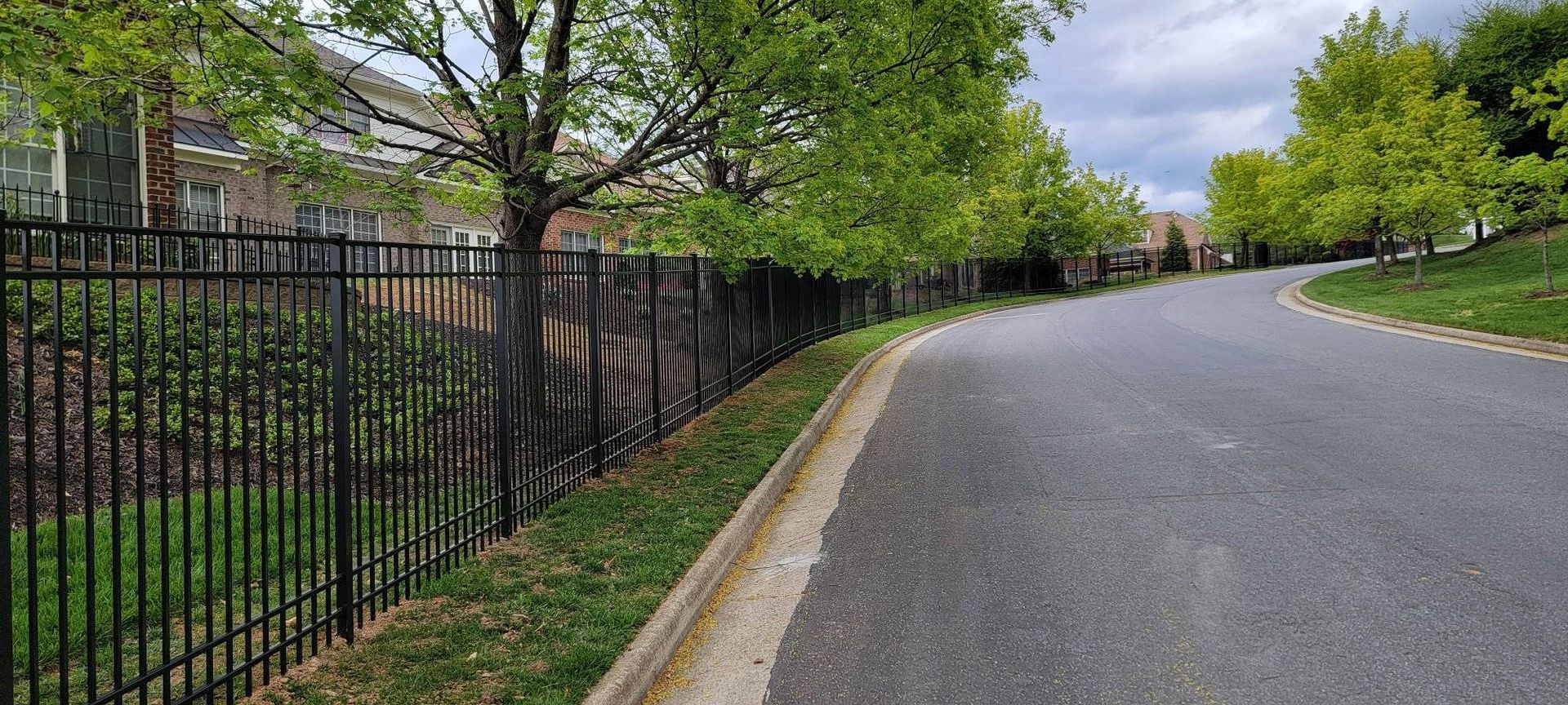 Black fence alongside a curving road with trees. Overcast sky.