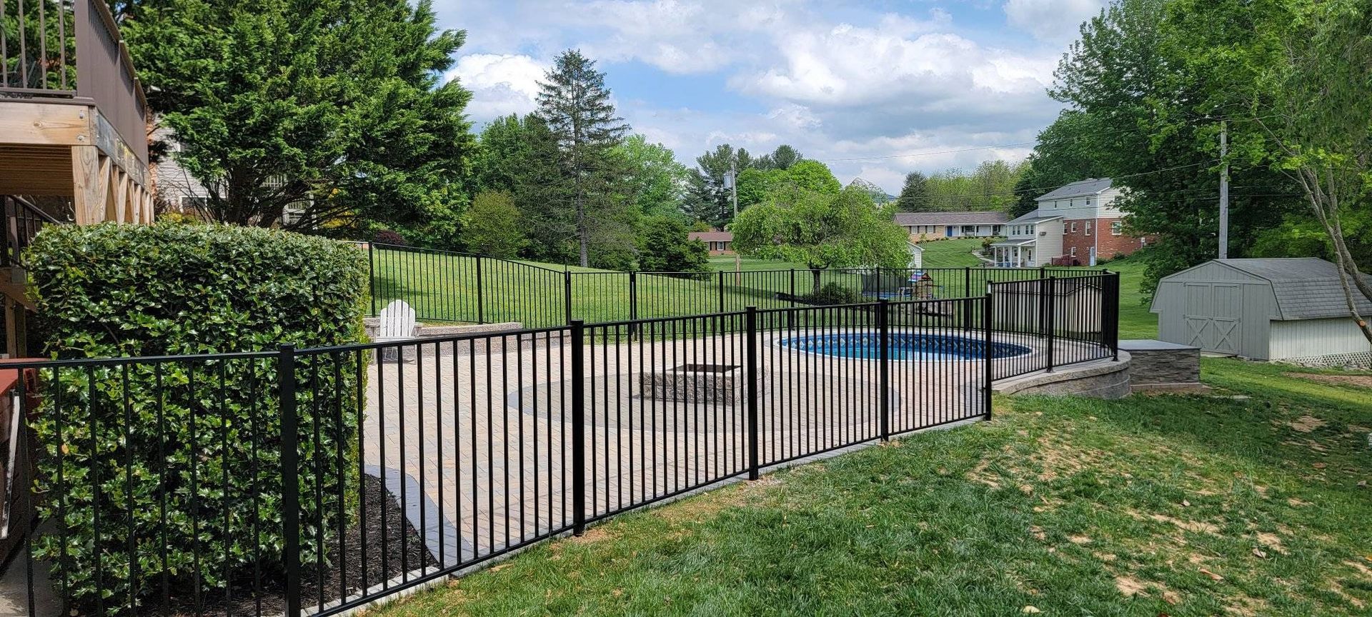 Black fenced-in swimming pool in a green yard, surrounded by trees and a building on a partly cloudy day.