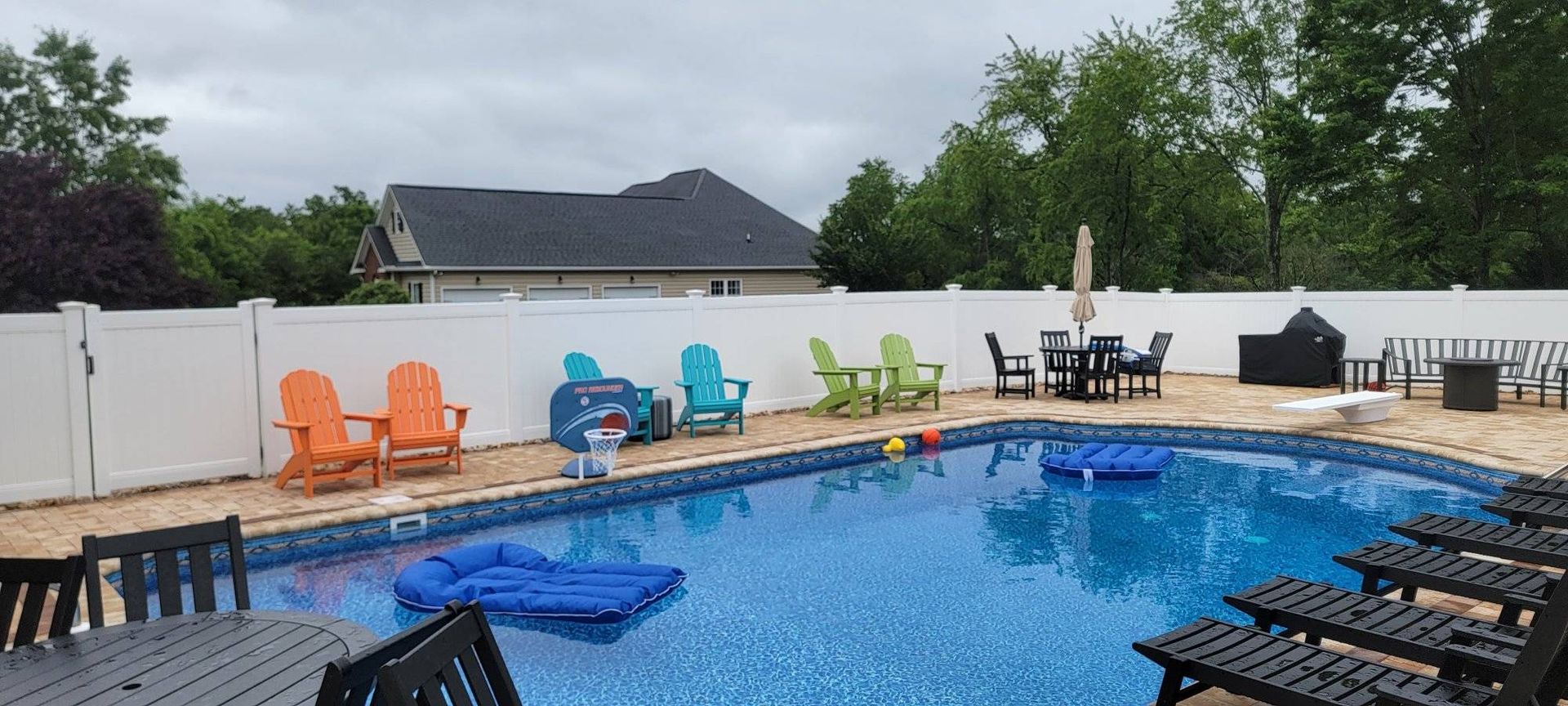 Backyard swimming pool with lounge chairs, tables, and colorful Adirondack chairs under an overcast sky.