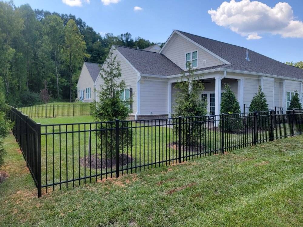 Black metal fence surrounding a light-colored house with a dark gray roof. Trees and grass are present.