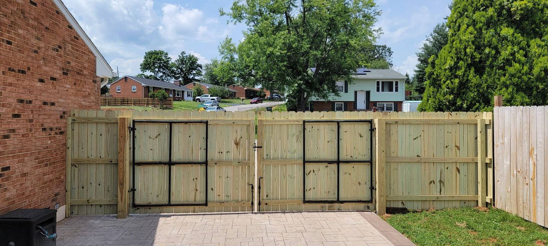 Wooden gates with black metal frames in a yard, brick wall on the left, grassy area and houses in the background.