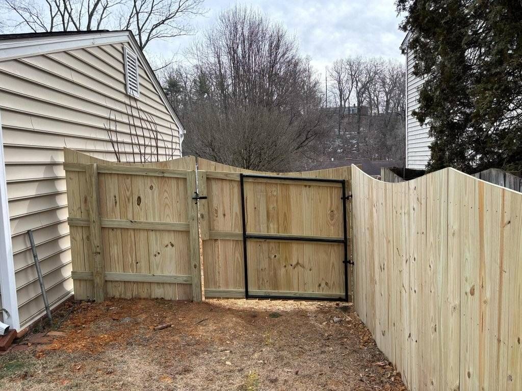 Wooden fence with gate, set against a beige-sided house, brown dirt, and trees in the background.