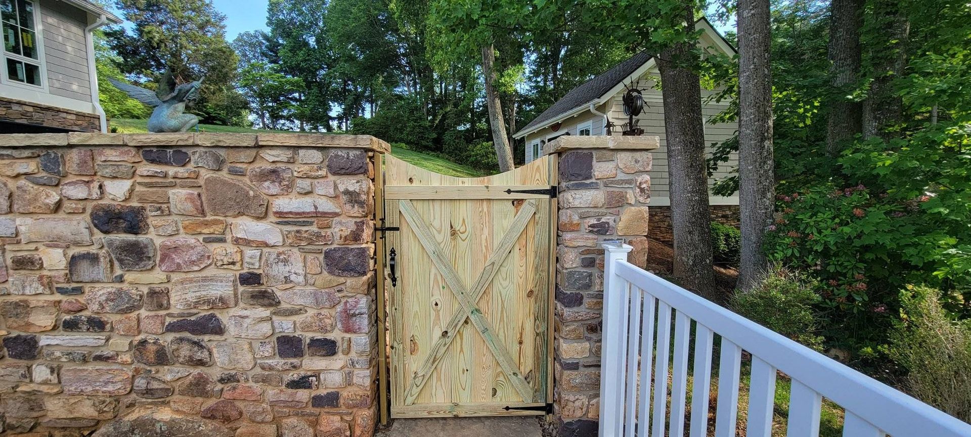Stone wall with wooden gate and white railing. Trees and a house are in the background.
