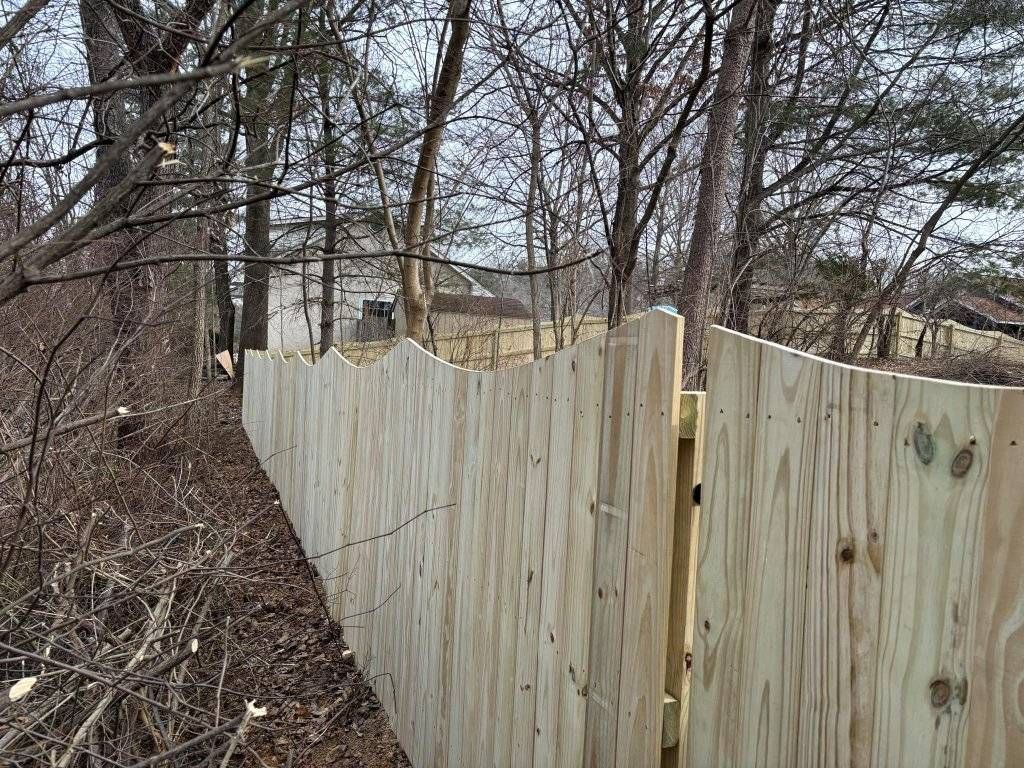 Wooden fence with a scalloped top, built in a wooded area. Trees in the background.