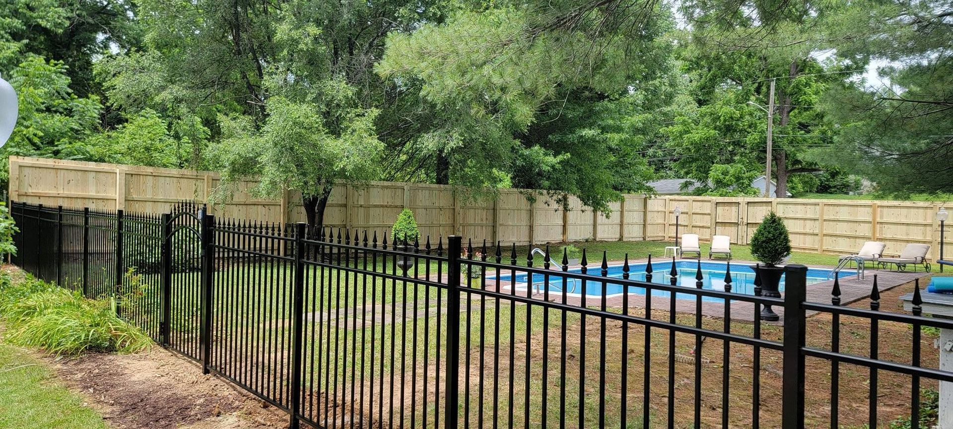 Black metal fence surrounds a swimming pool, beyond a wooden fence and trees.