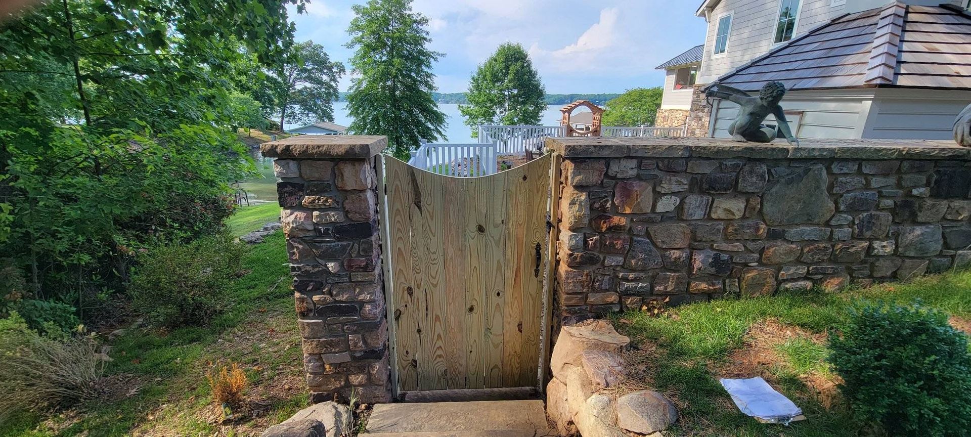 Stone gate with wooden door, leading to a lake. Trees and a house visible in the background.
