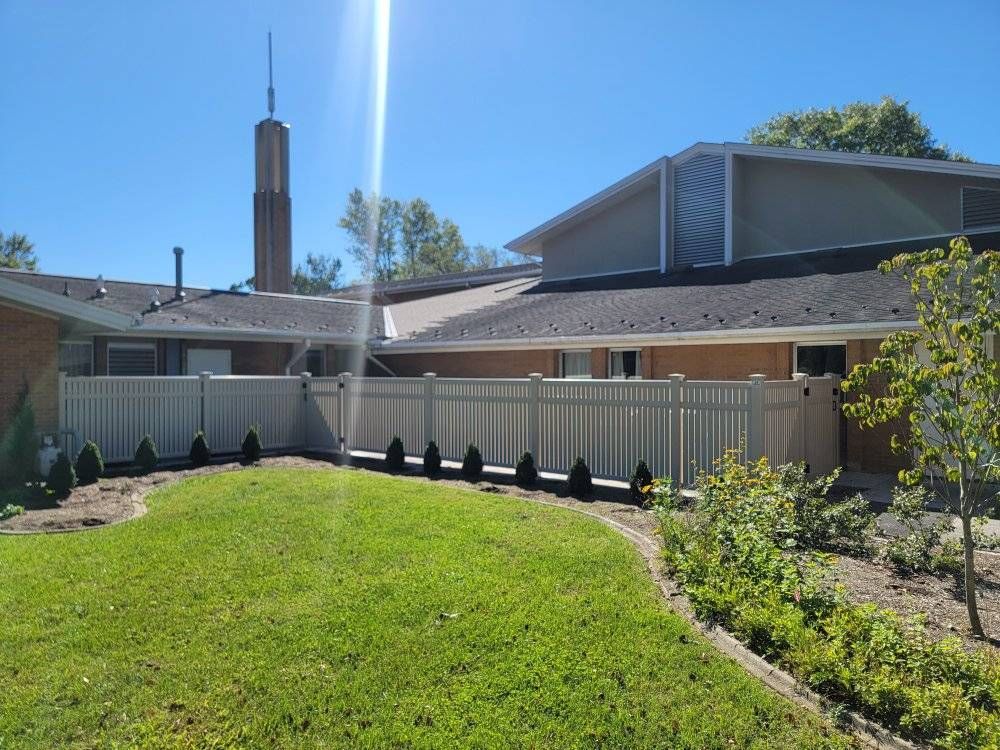 A tan fence encloses a green lawn and building with a brown roof under a sunny, blue sky.