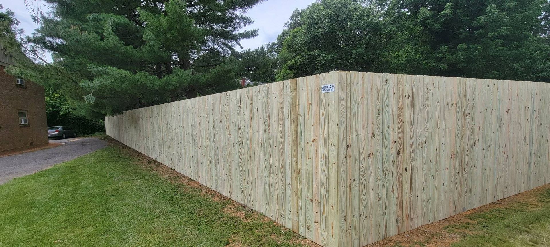 A wooden fence bordering a lawn. Tall trees are behind the fence under a cloudy sky.