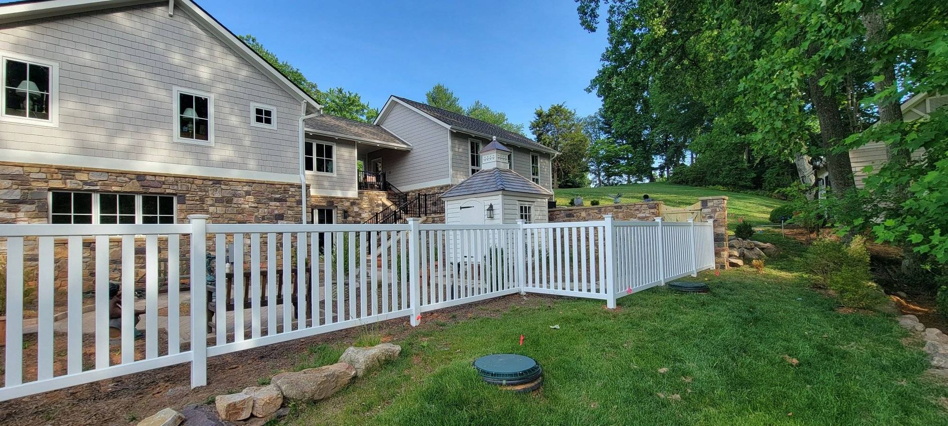 A house behind a white picket fence on a grassy hill. Trees on the right.