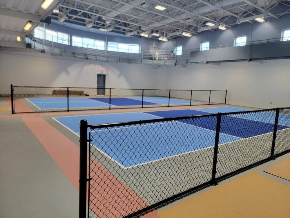 Indoor pickleball courts with blue and grey surfaces, enclosed by black fencing, in a brightly lit gym.