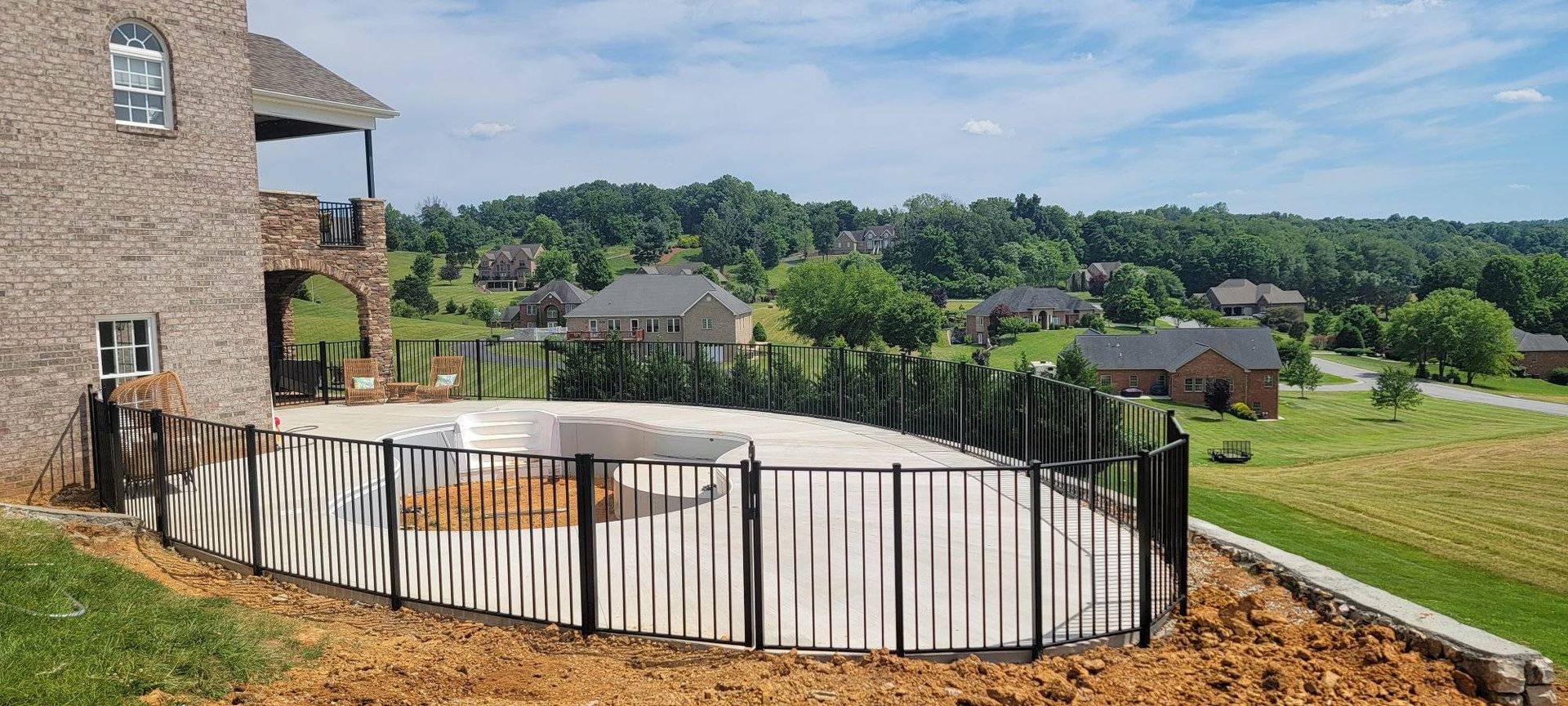A partially built swimming pool surrounded by a black fence, next to a stone house and overlooking a green landscape.