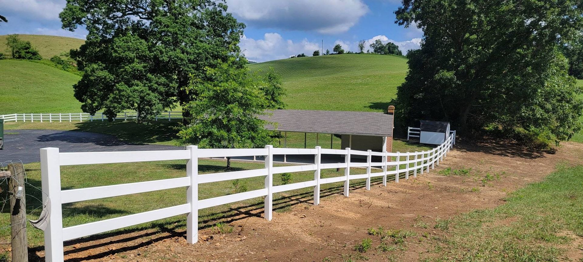 A white fence curves around a grassy area with trees and a building, rolling green hills in the background.