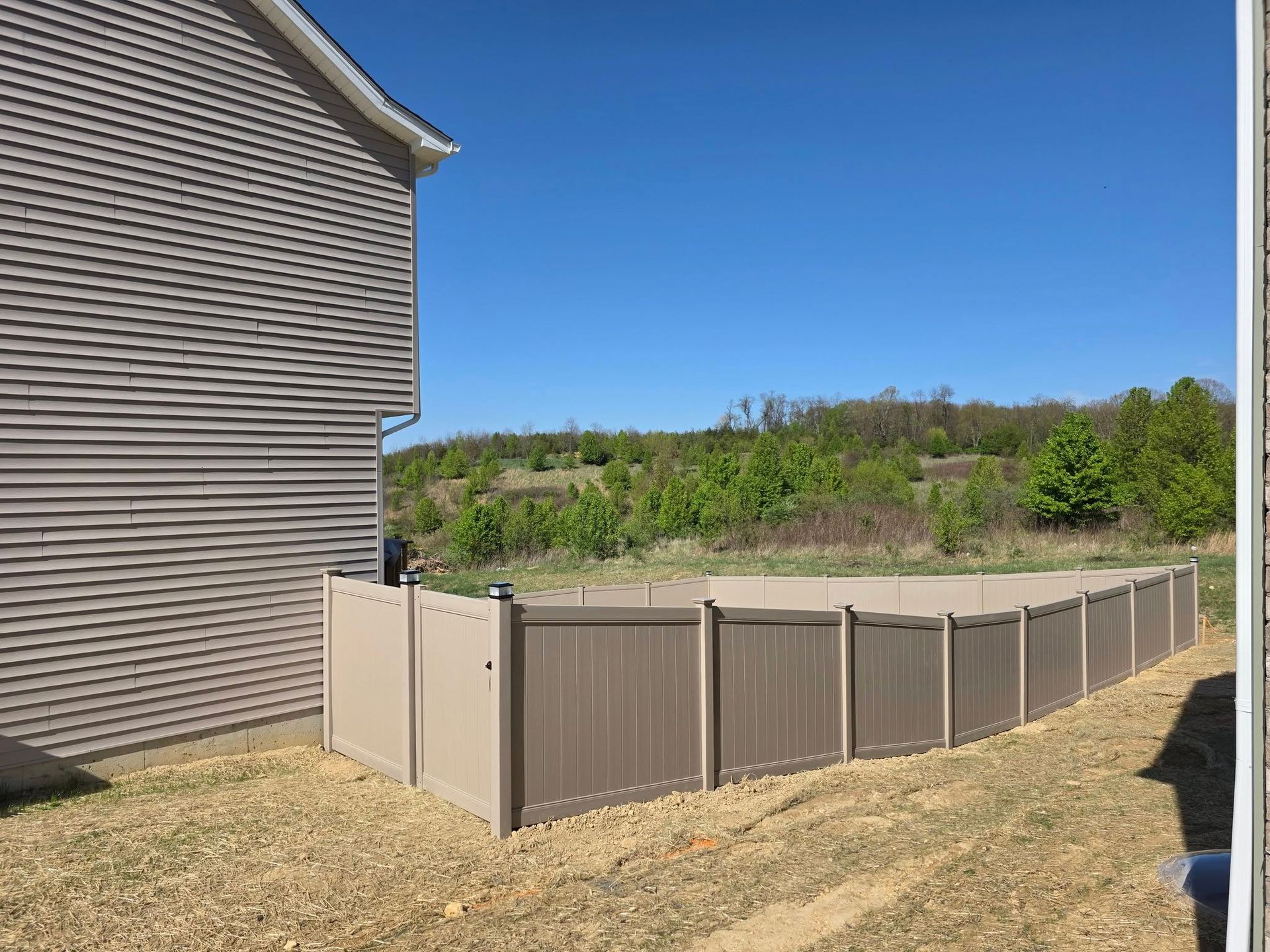 Tan vinyl fence surrounds a backyard near a house. Hills and blue sky in background.