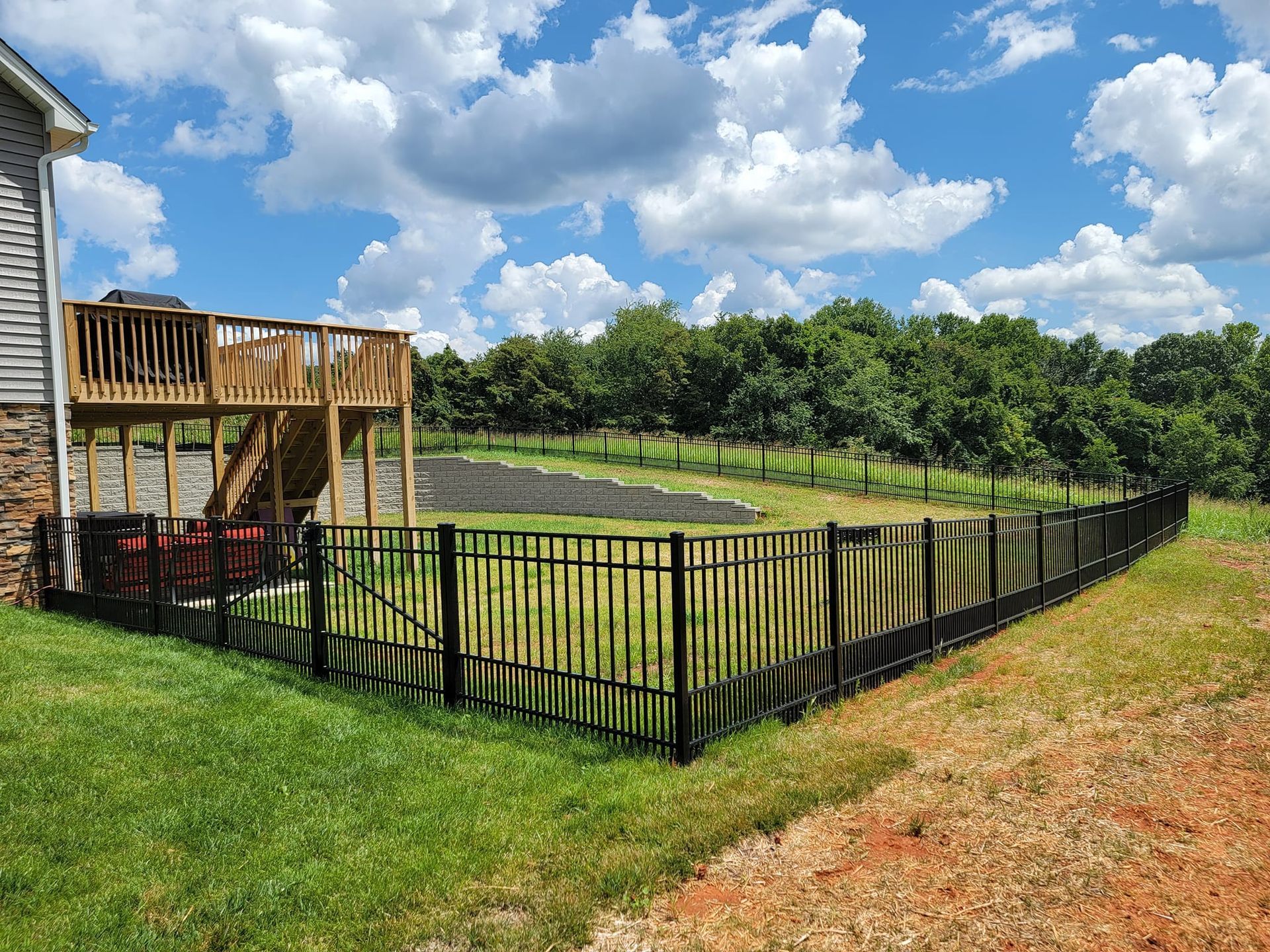 Black fence encloses backyard, deck attached to house. Green grass and trees under a blue sky.