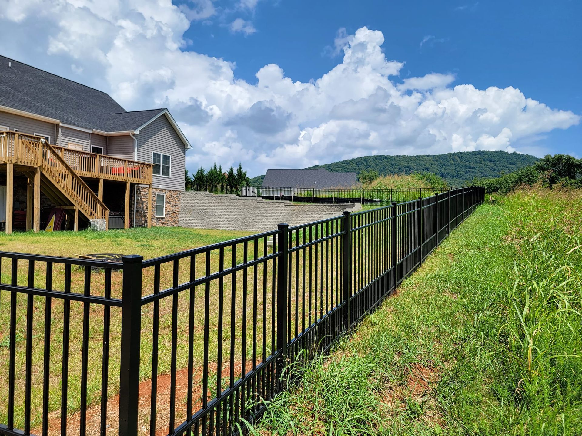 Black metal fence bordering a grassy area next to a house with a wooden deck; mountain in the background.