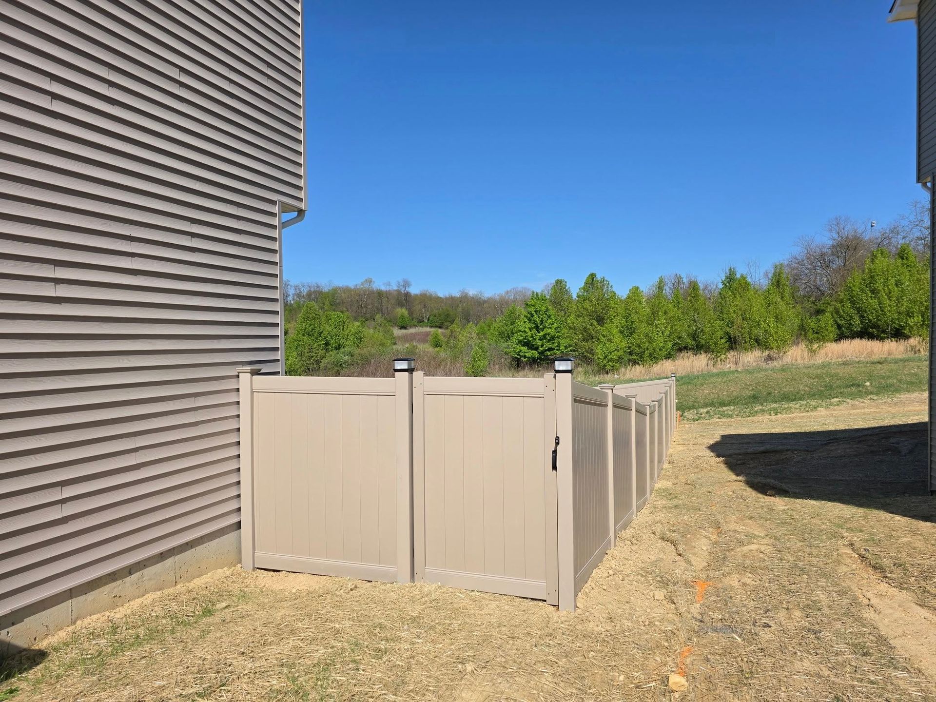 Tan vinyl fence enclosing the side of a beige-sided house, grassy yard, trees in the background, blue sky.