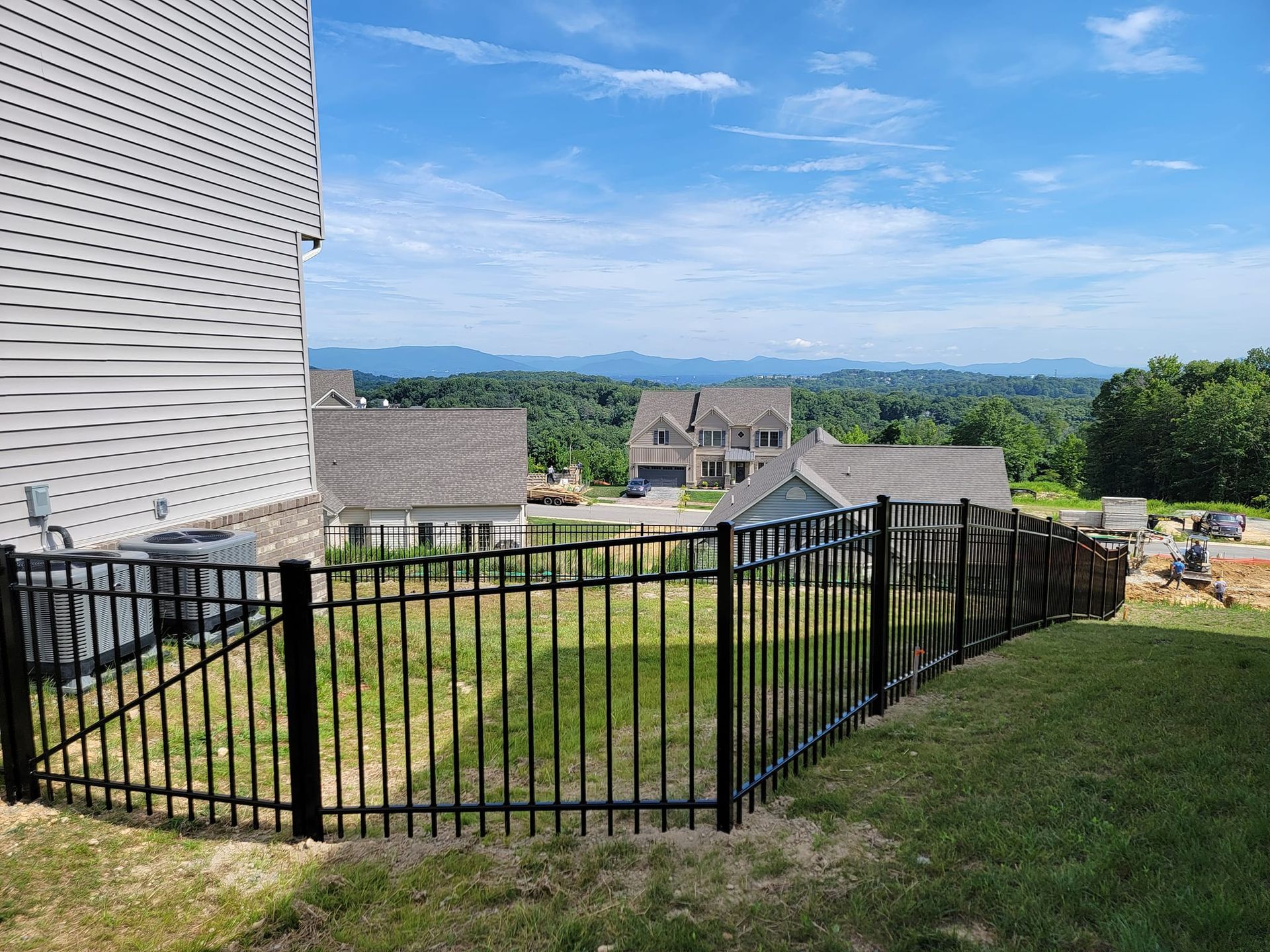 Black metal fence curves along a grassy yard, with houses and distant mountains visible under a blue sky.