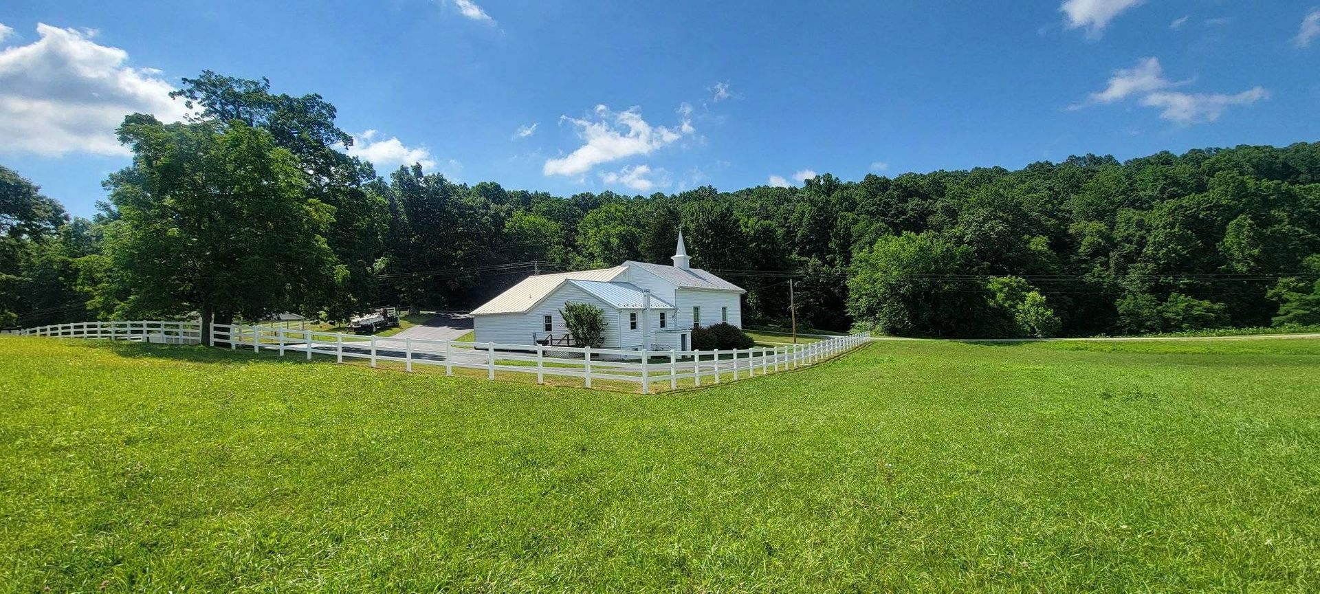 White building with a fence in a green field, forest in the background, blue sky with clouds.