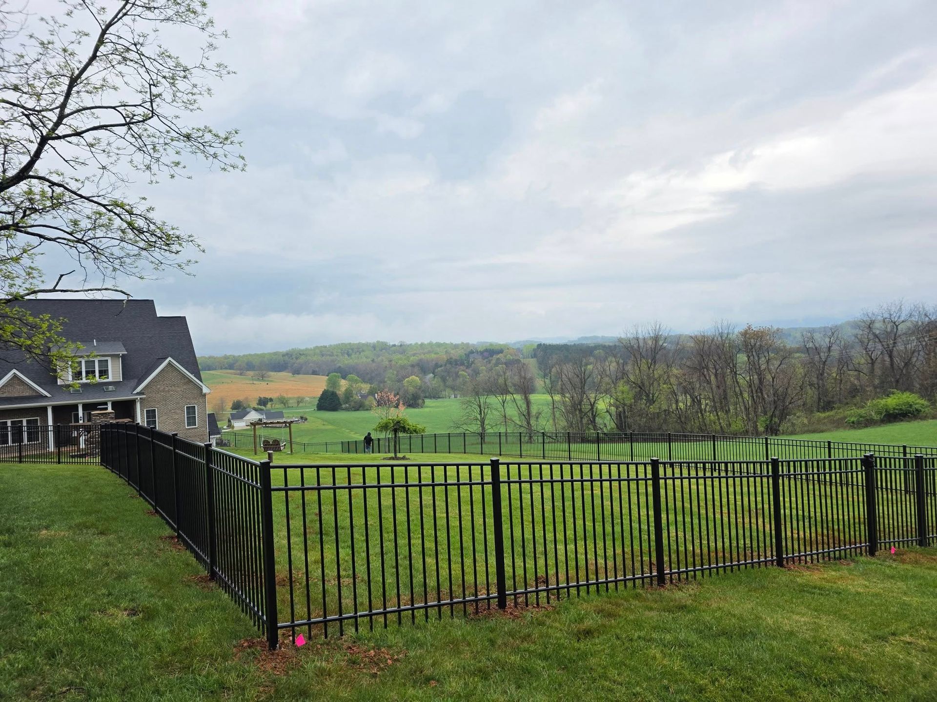 Black metal fence enclosing a grassy yard overlooking a valley under a cloudy sky.