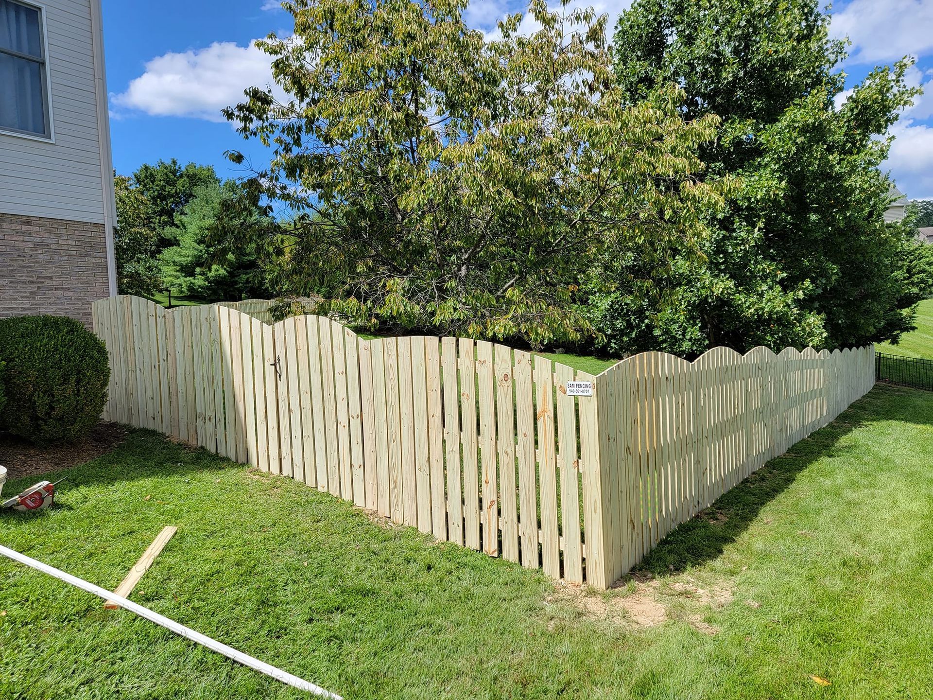 Wooden fence in a yard with green grass, shrubs, and trees against a blue sky.