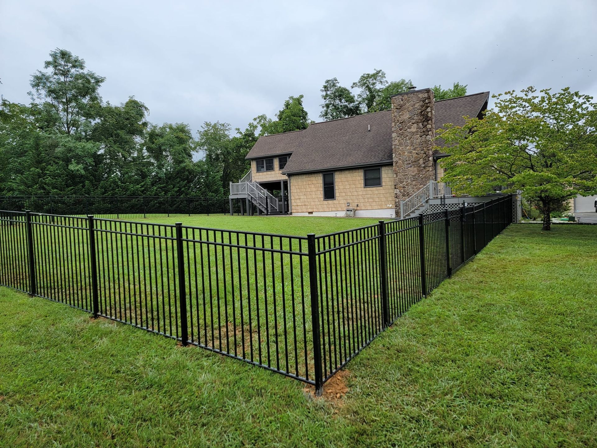 Black metal fence surrounds a grassy yard, with a brick house and trees in the background. Overcast sky.