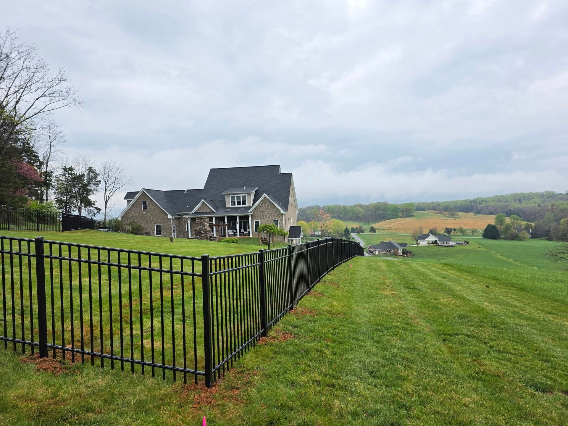 Black fence bordering grassy hill, large house in the background, cloudy sky.