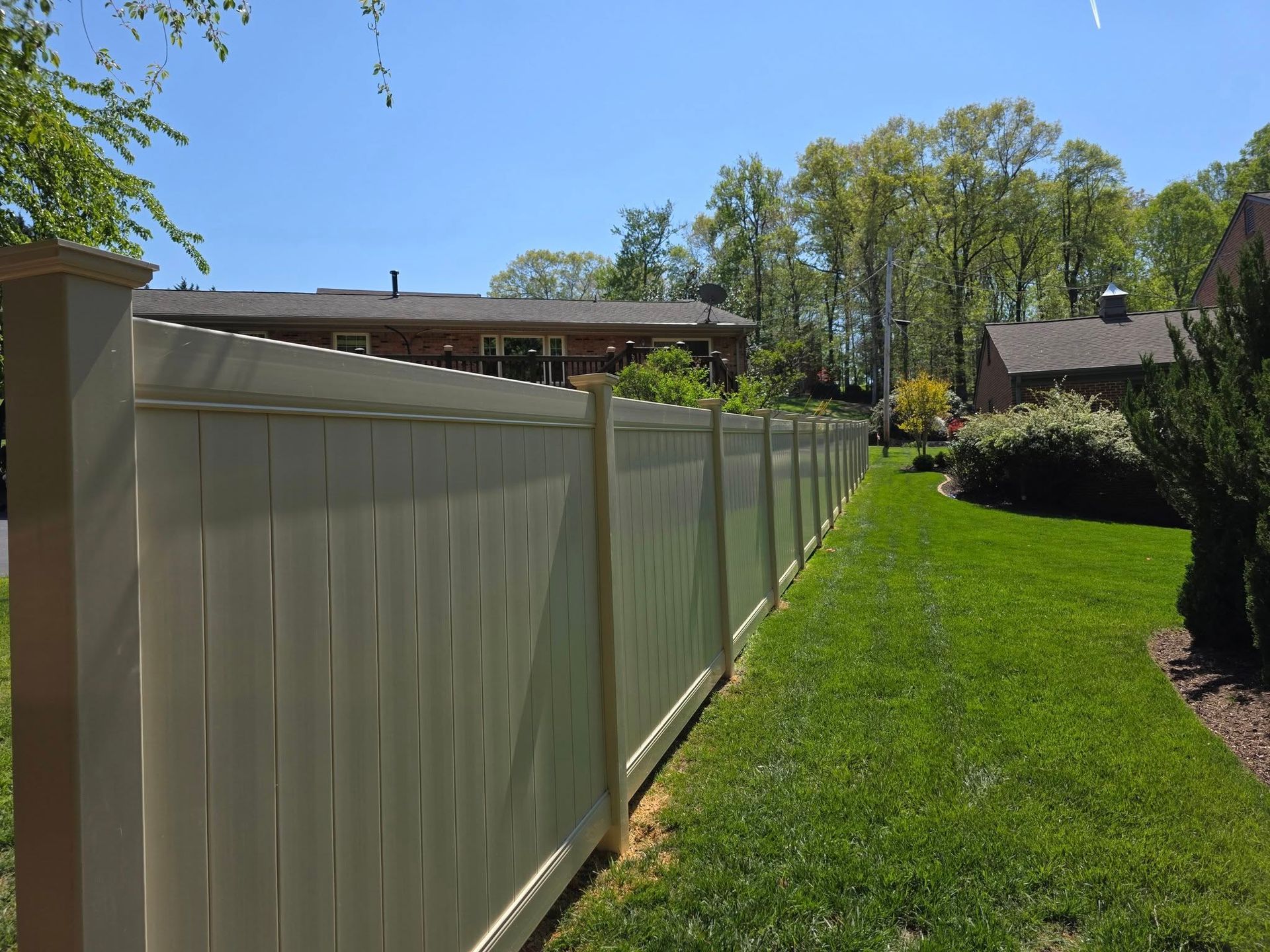 Cream-colored privacy fence along a green lawn, with houses and trees in the background under a blue sky.