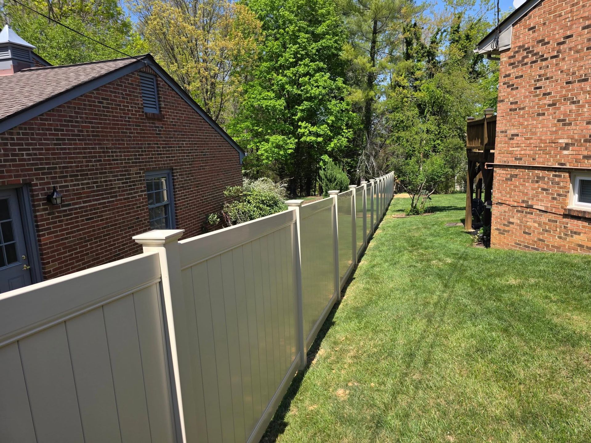 Beige fence runs along a green grassy yard between brick buildings and trees on a sunny day.