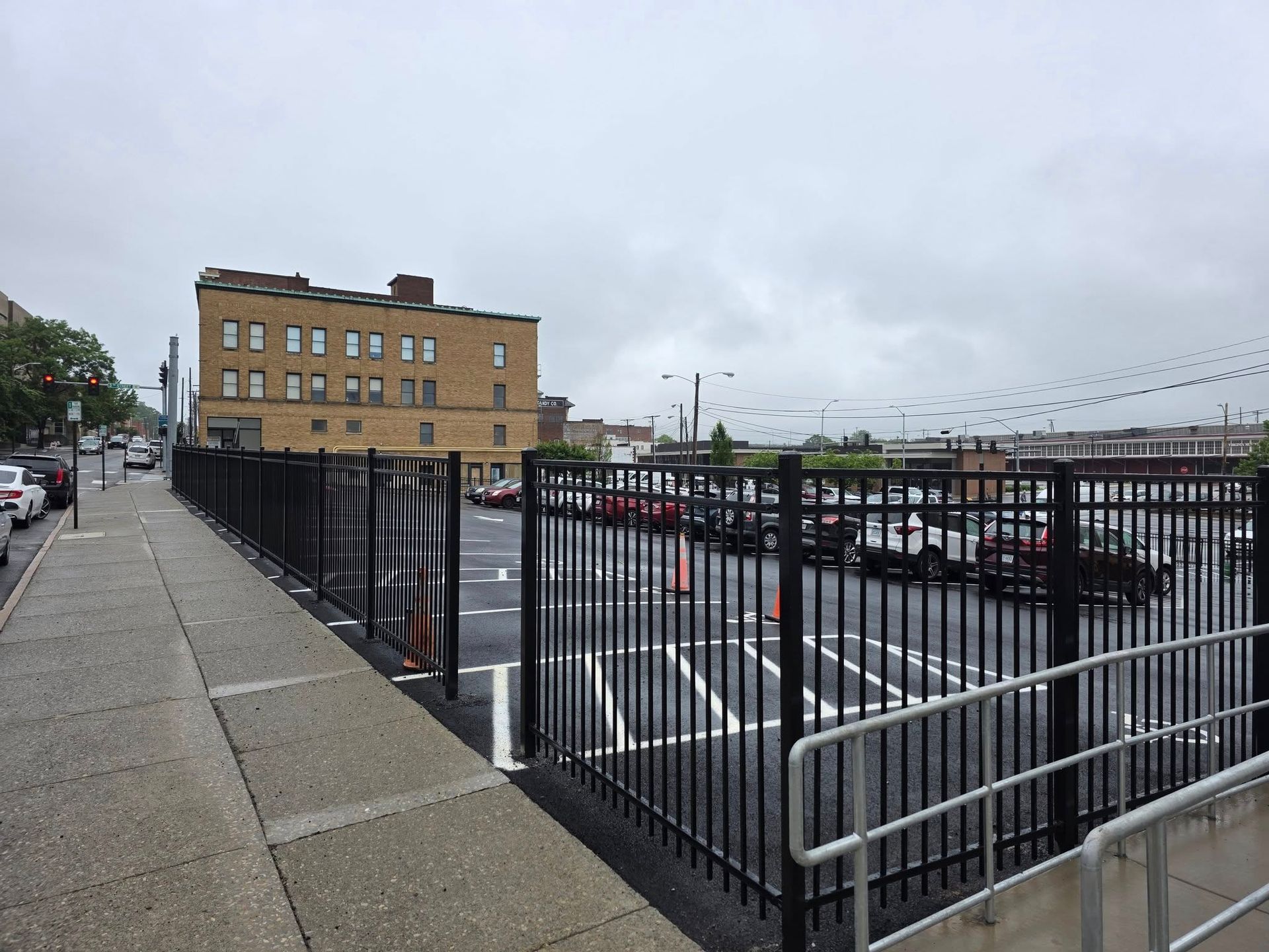 Fenced parking lot next to a sidewalk and street, with a brick building in the background under an overcast sky.