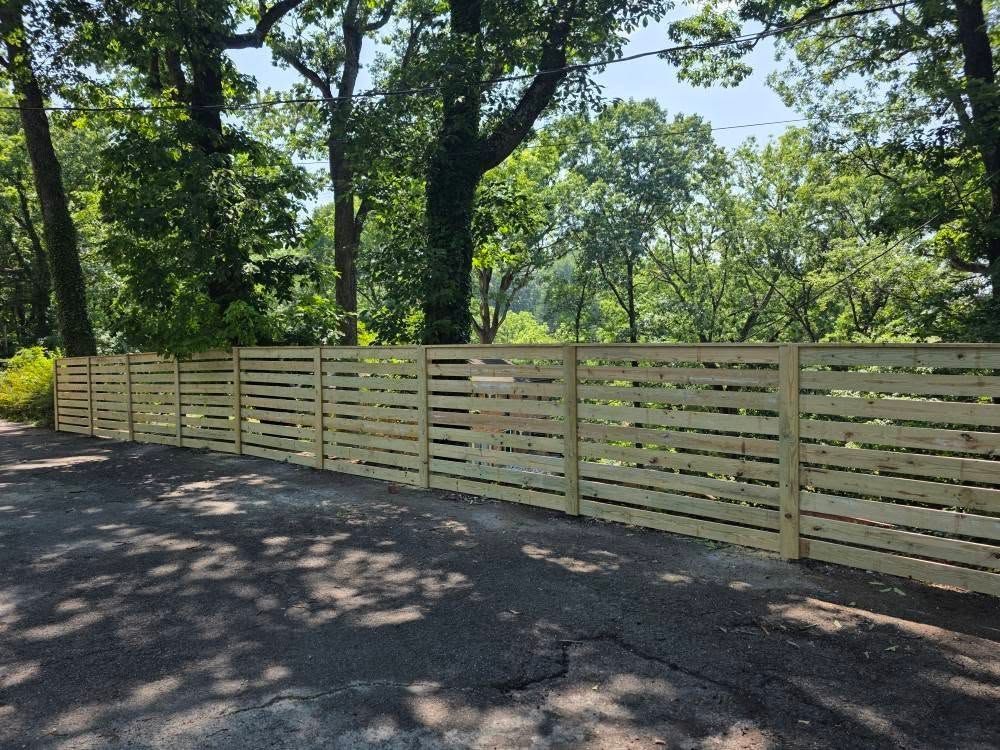 A light-colored horizontal slat wooden fence along a paved road, with trees in the background.