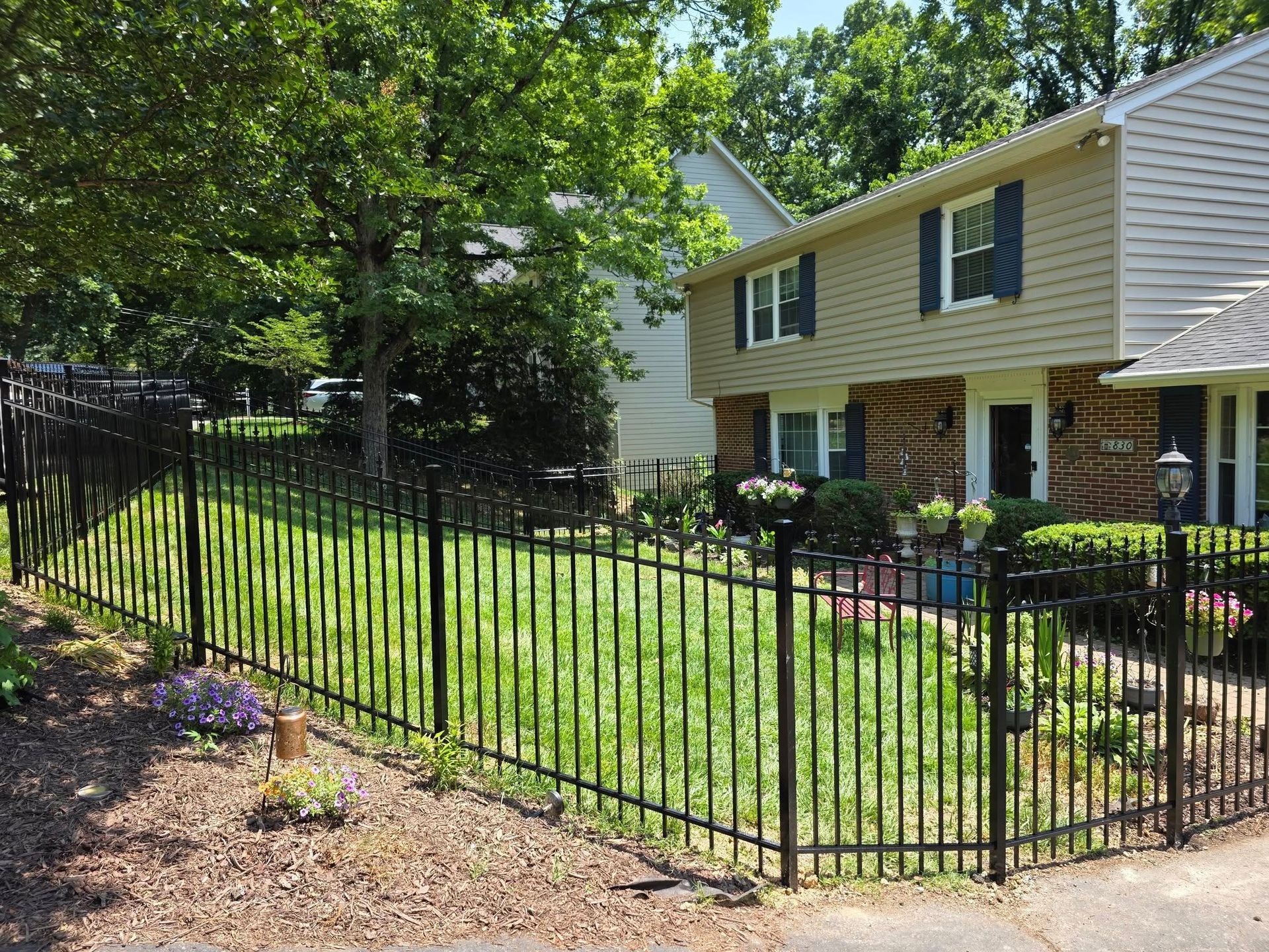 Black metal fence encloses a green lawn in front of a two-story house with tan siding and dark shutters.