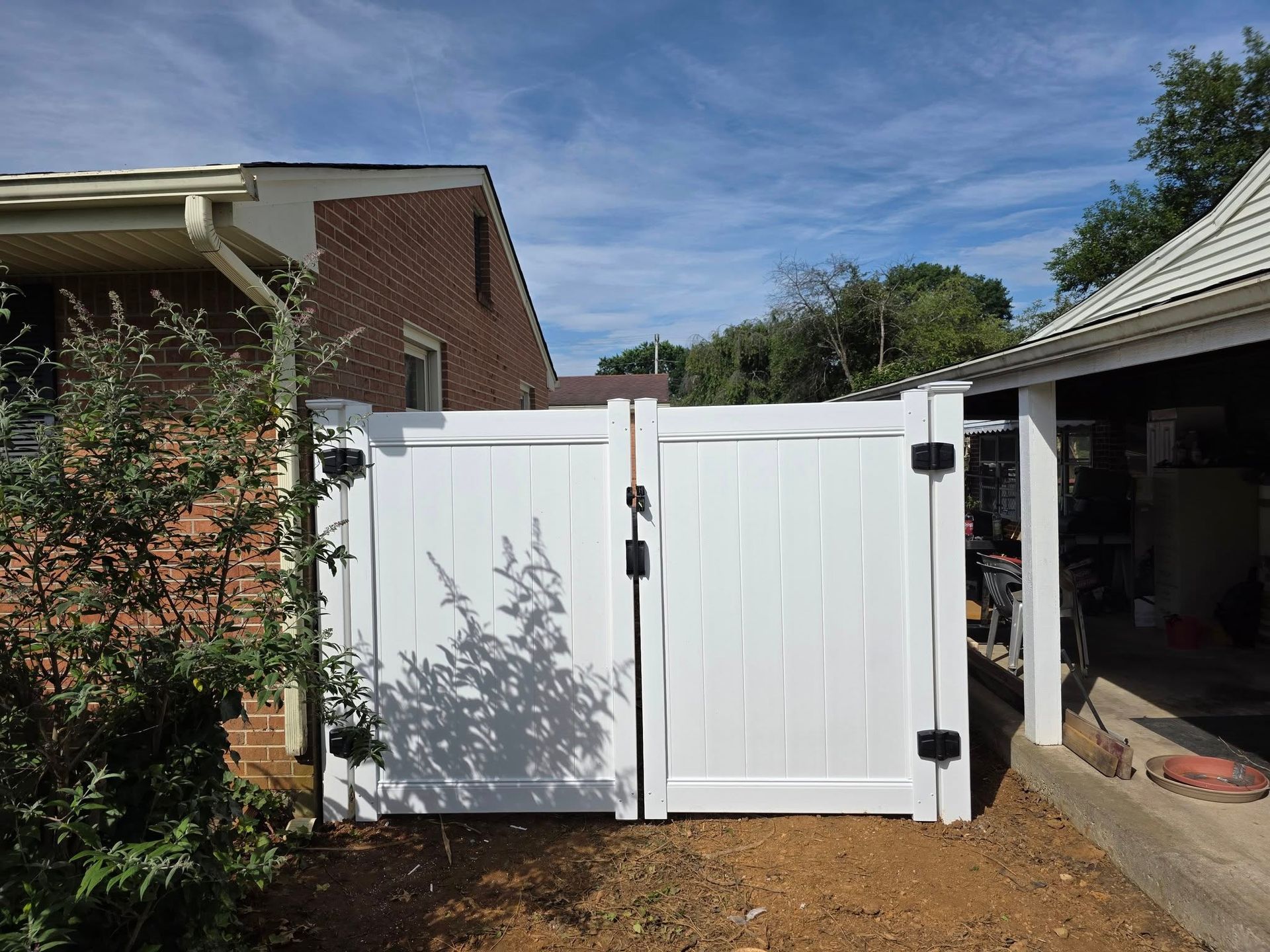 White double gate with black hinges and latch, between a brick house and a white-roofed structure under a blue sky.