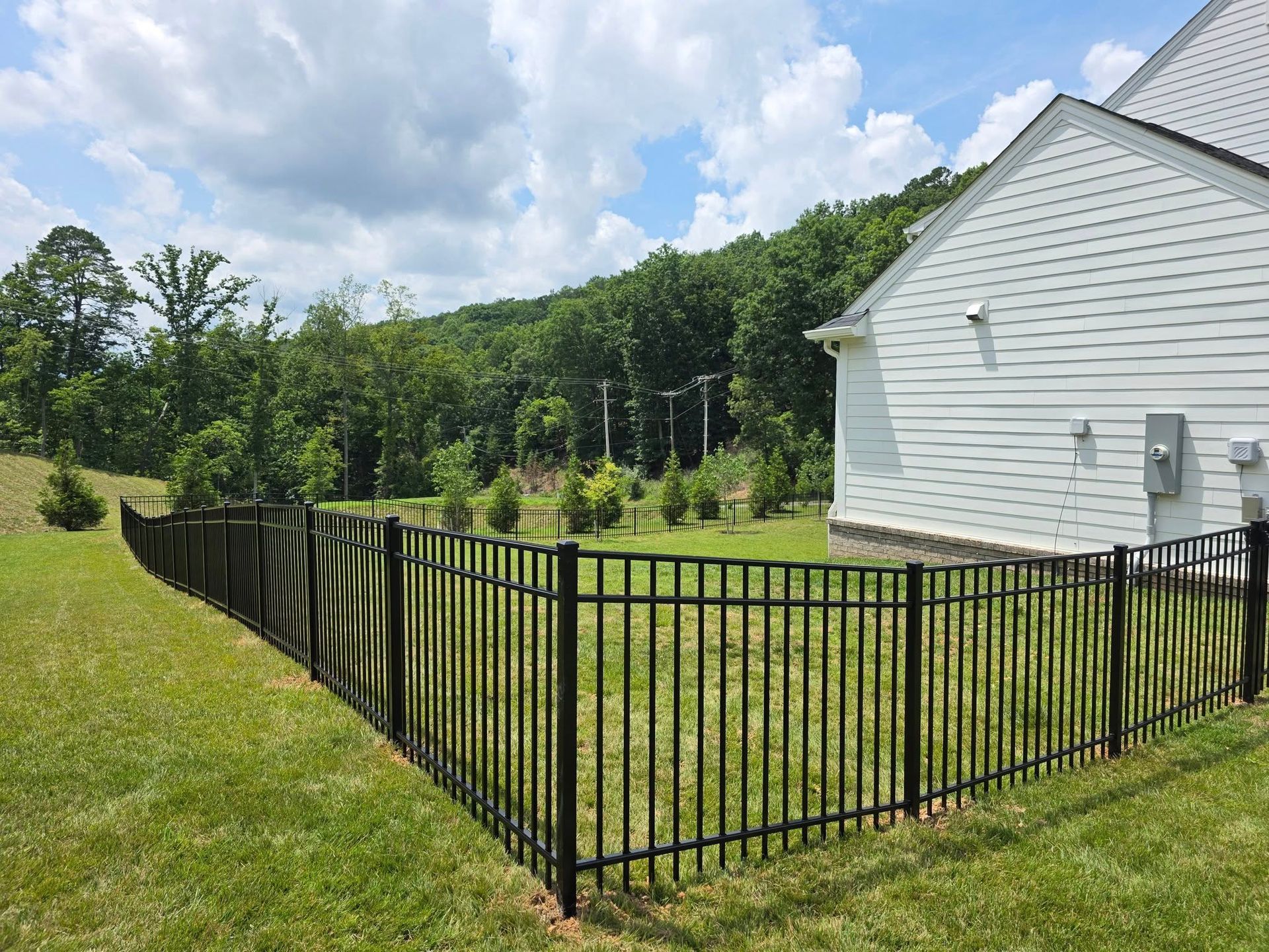 Black metal fence surrounds grassy yard next to a white house with a wooded hillside in the background under a cloudy sky.