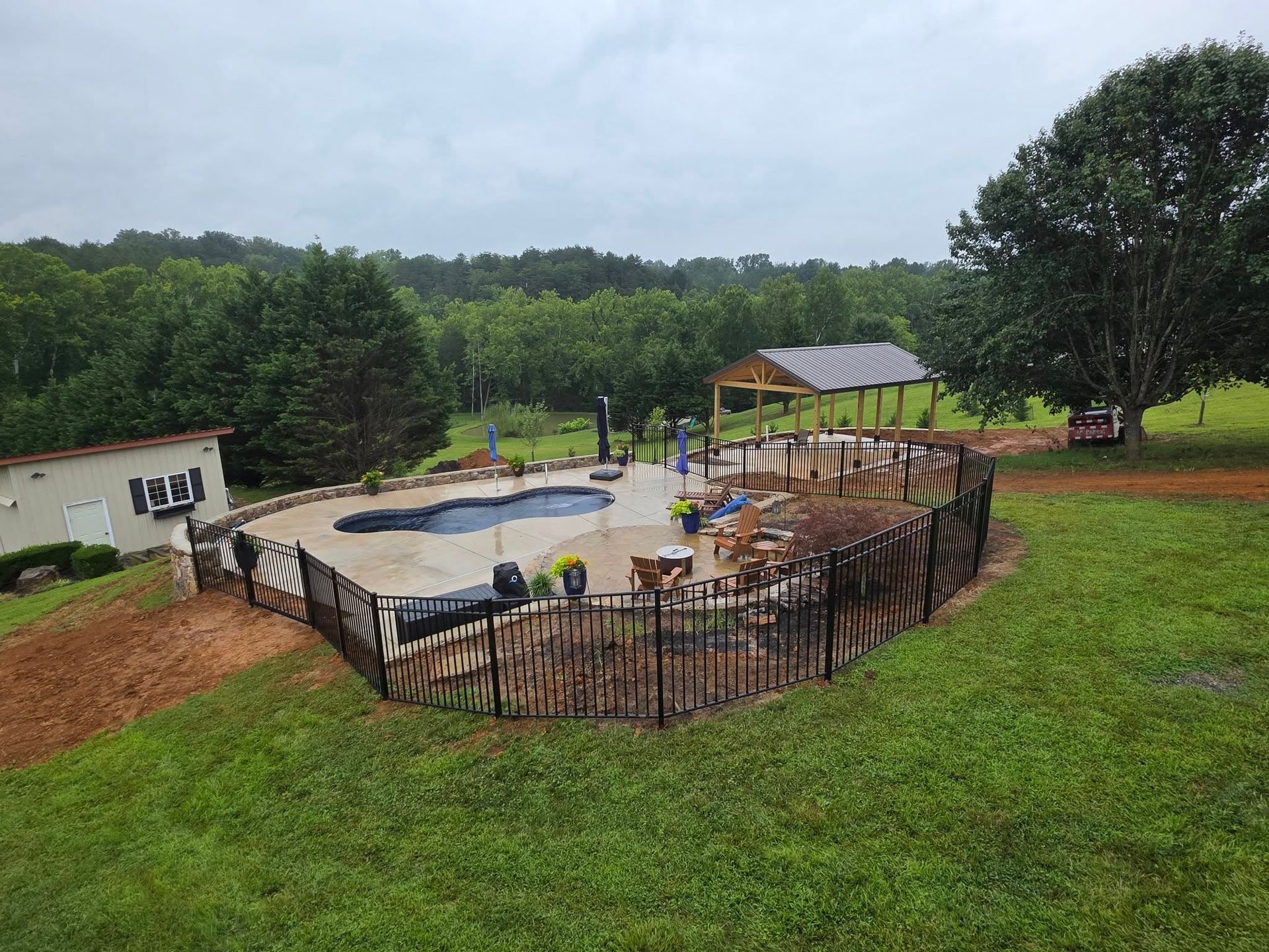 Pool area with dark fence, surrounded by green grass and trees. Cloudy sky.