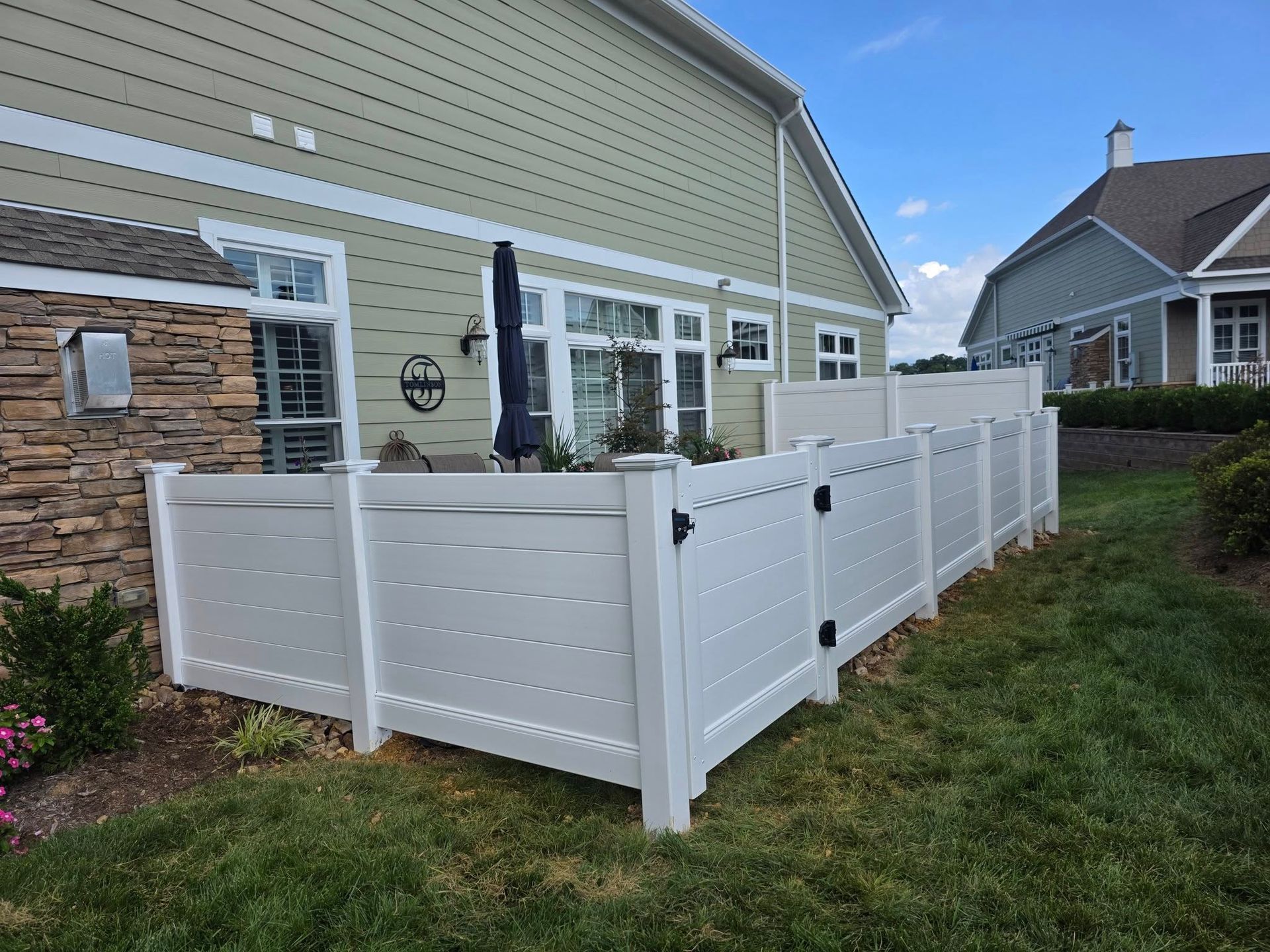 White vinyl fence encloses a patio next to a light green house with a blue umbrella.