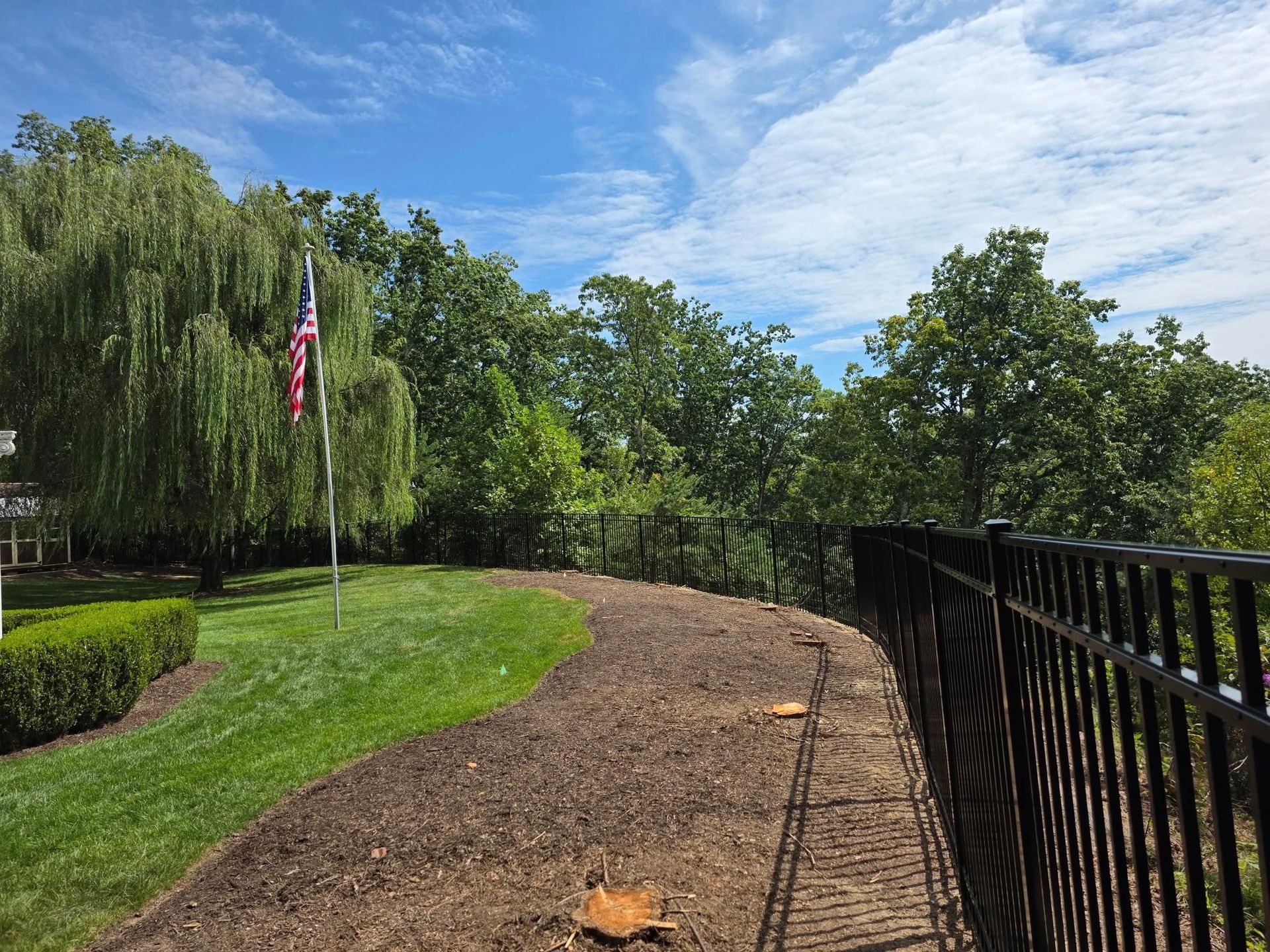 A curved pathway with fresh mulch and a black metal fence, bordered by green grass and trees, and an American flag on a sunny day.