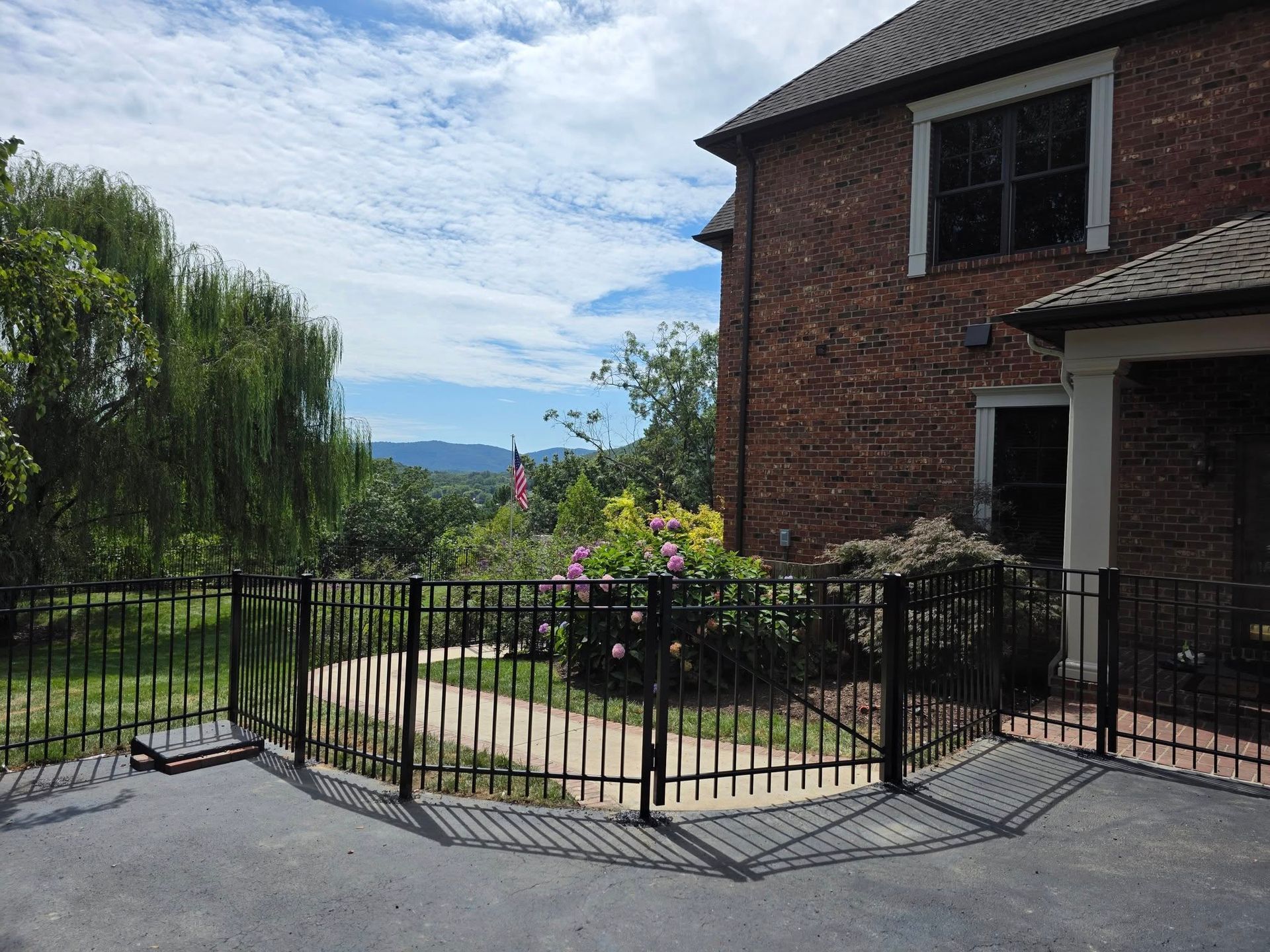Black fence surrounds a brick house with a view of green trees and distant mountains under a cloudy sky.