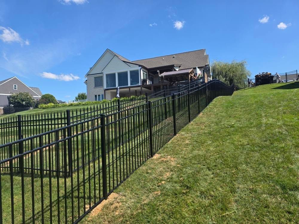 Black fence along a grassy slope, with a two-story house under a blue sky in the background.