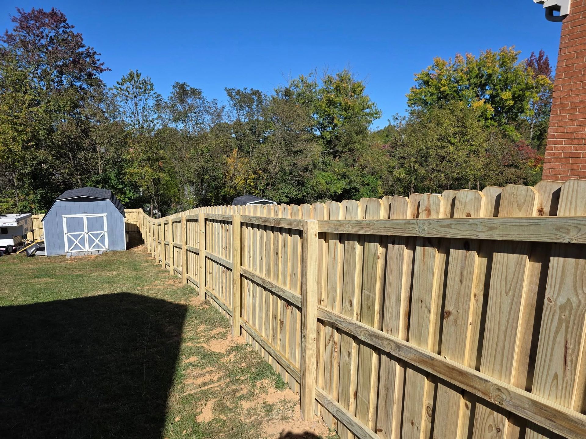 Wooden fence surrounds a yard with a blue shed, trees, and a brick building under a clear sky.